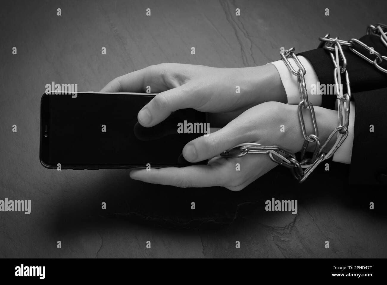 Closeup view of internet addicted woman with chained hands using smartphone at dark table, space for text. Black and white effect Stock Photo