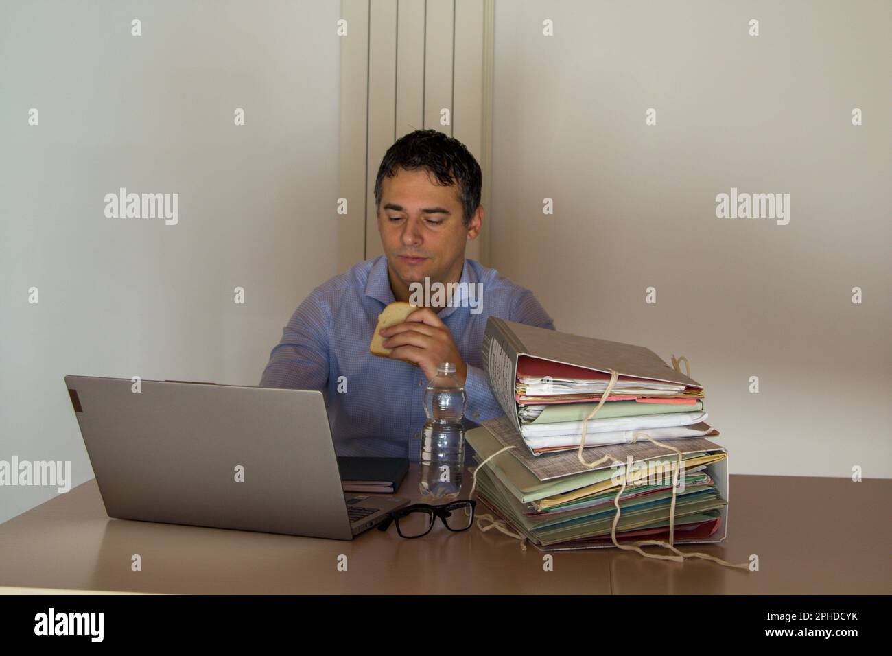 Image of a desperate man holding a sandwich and a small bottle of water who has to work overtime in his office due to overwork. Stock Photo