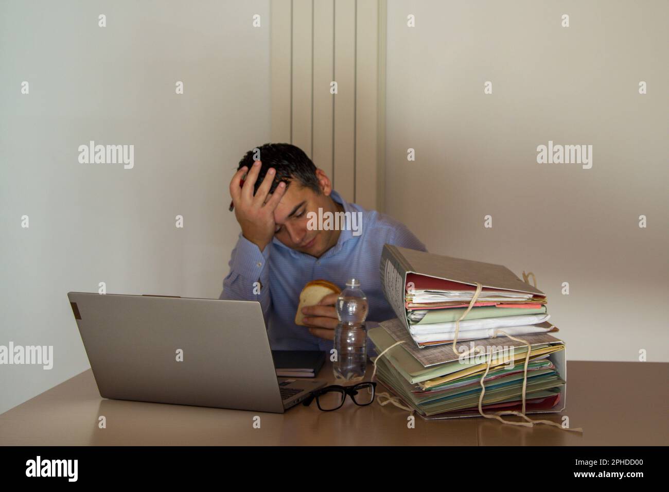 Image of a desperate man holding a sandwich and a small bottle of water who has to work overtime in his office due to overwork. Stock Photo