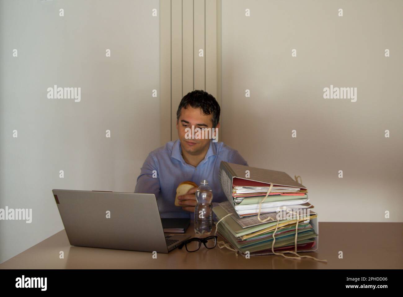 Image of a desperate man holding a sandwich and a small bottle of water who has to work overtime in his office due to overwork. Stock Photo