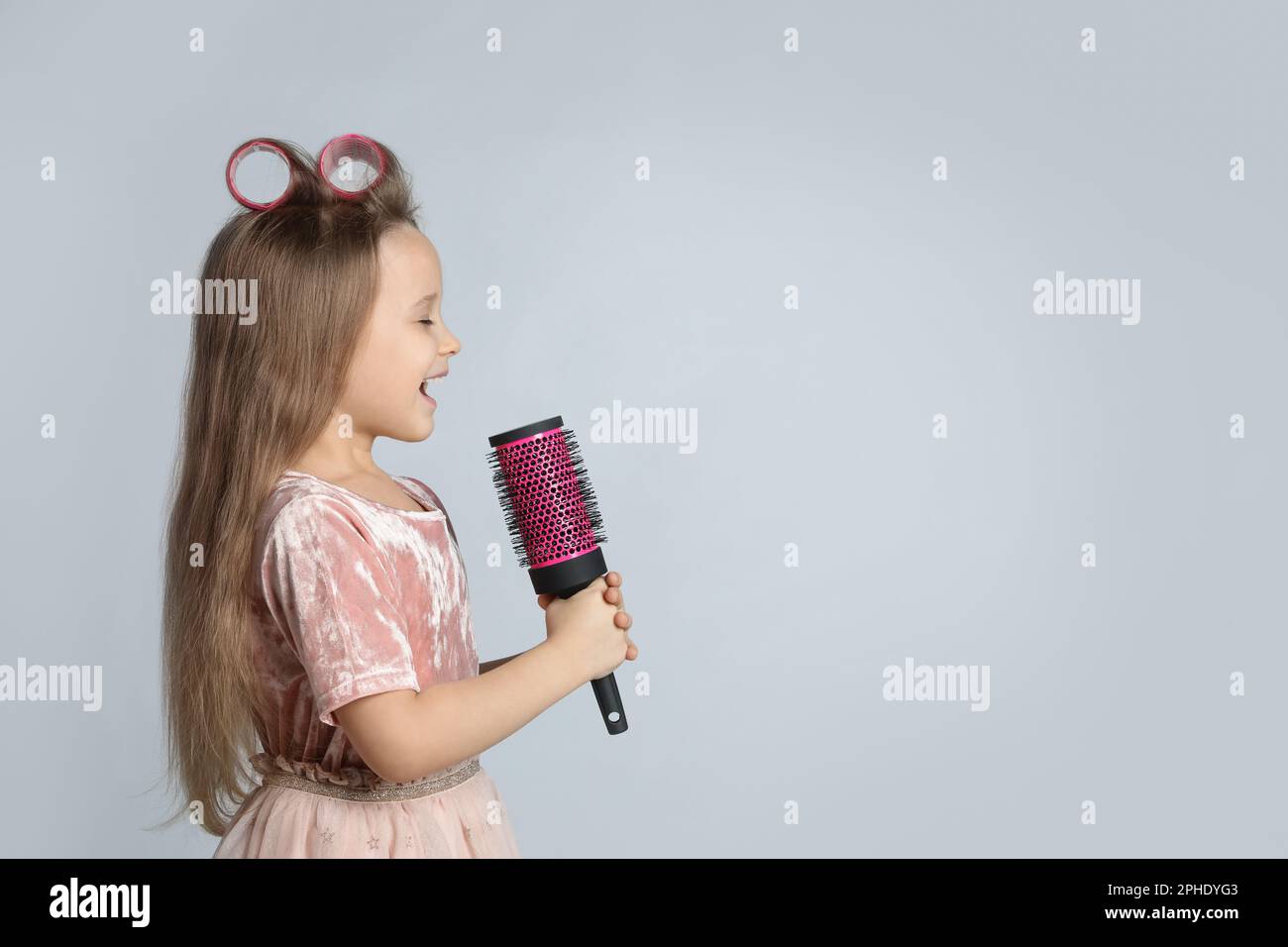 Cute little girl with hairbrush singing on light grey background, space