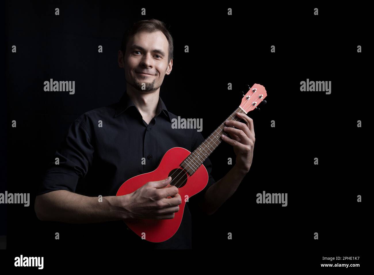 Handsome smiling low key man portrait playing on fun red ukulele guitar over black background Stock Photo