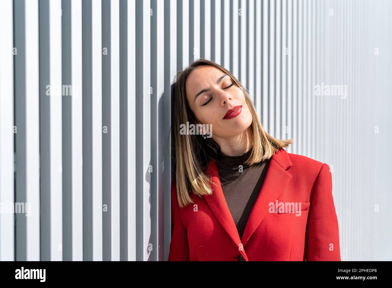 Young female in red coat and dark blonde hair leaning on gray wall with closed eyes in sunny day Stock Photo