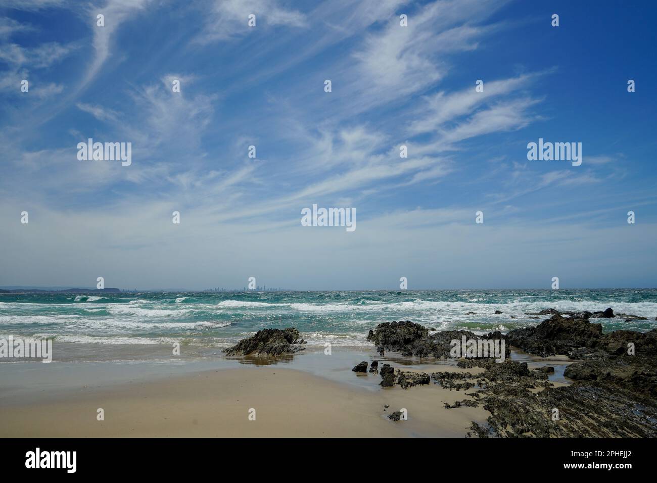 Surf beach on a windy day. View from sandy shore over rocks and surf to city skyline on the horizon, with windswept cirrus clouds against a blue sky. Stock Photo