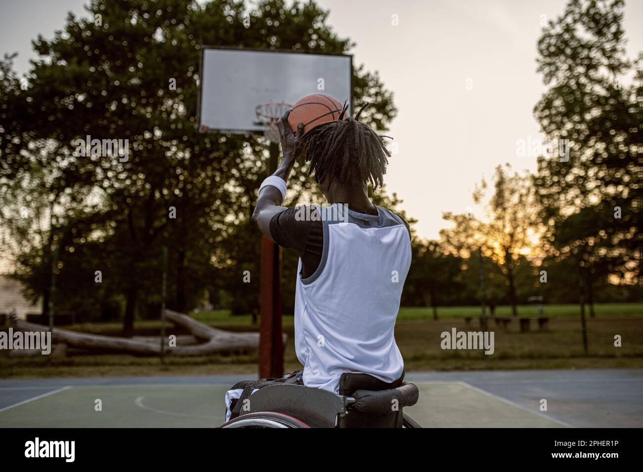 Young athlete on wheelchair throwing ball in basketball basket, sunset