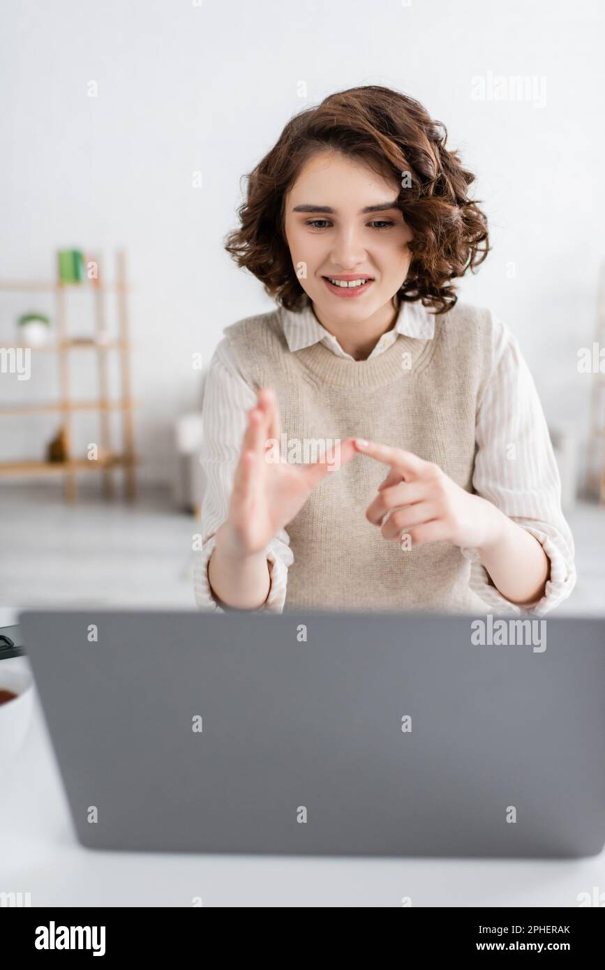 cheerful woman with curly hair teaching sign language alphabet near blurred laptop at home,stock