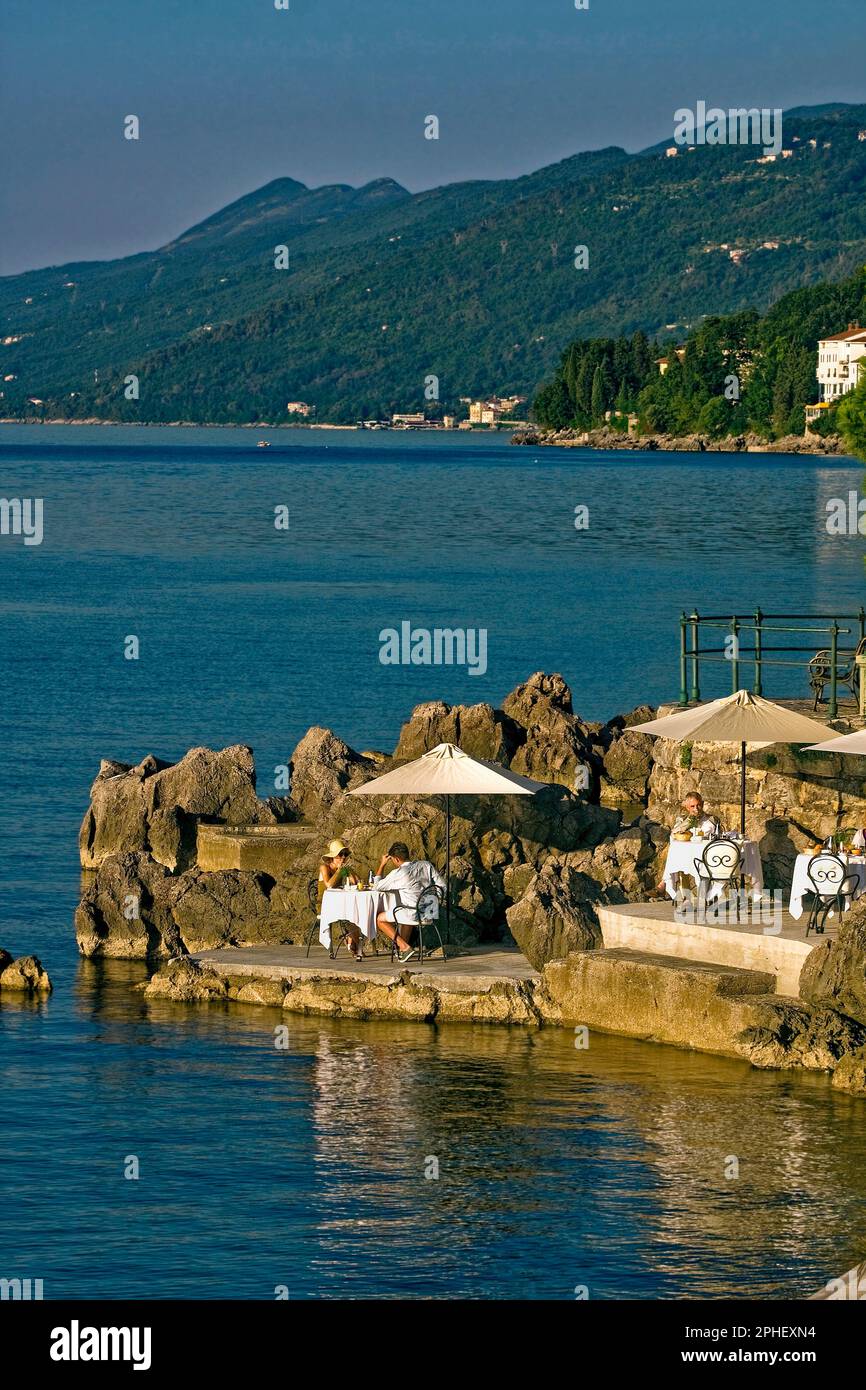 waterfront scene; people eating, tables, umbrellas, rocky prominence, peaceful, early morning,golden light, coastal, hills beyond, water, Lungomare; p Stock Photo