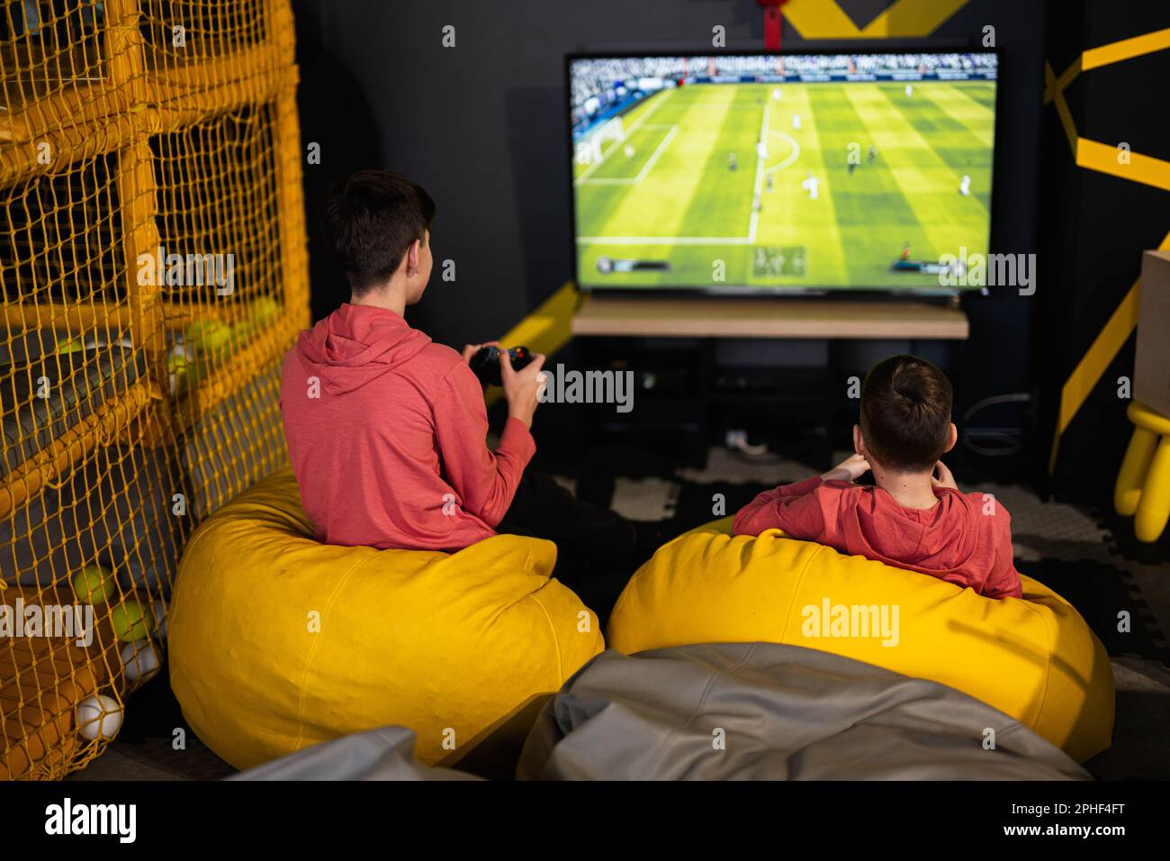 Two brothers playing football video game console, sitting on yellow pouf in  kids play center Stock Photo - Alamy