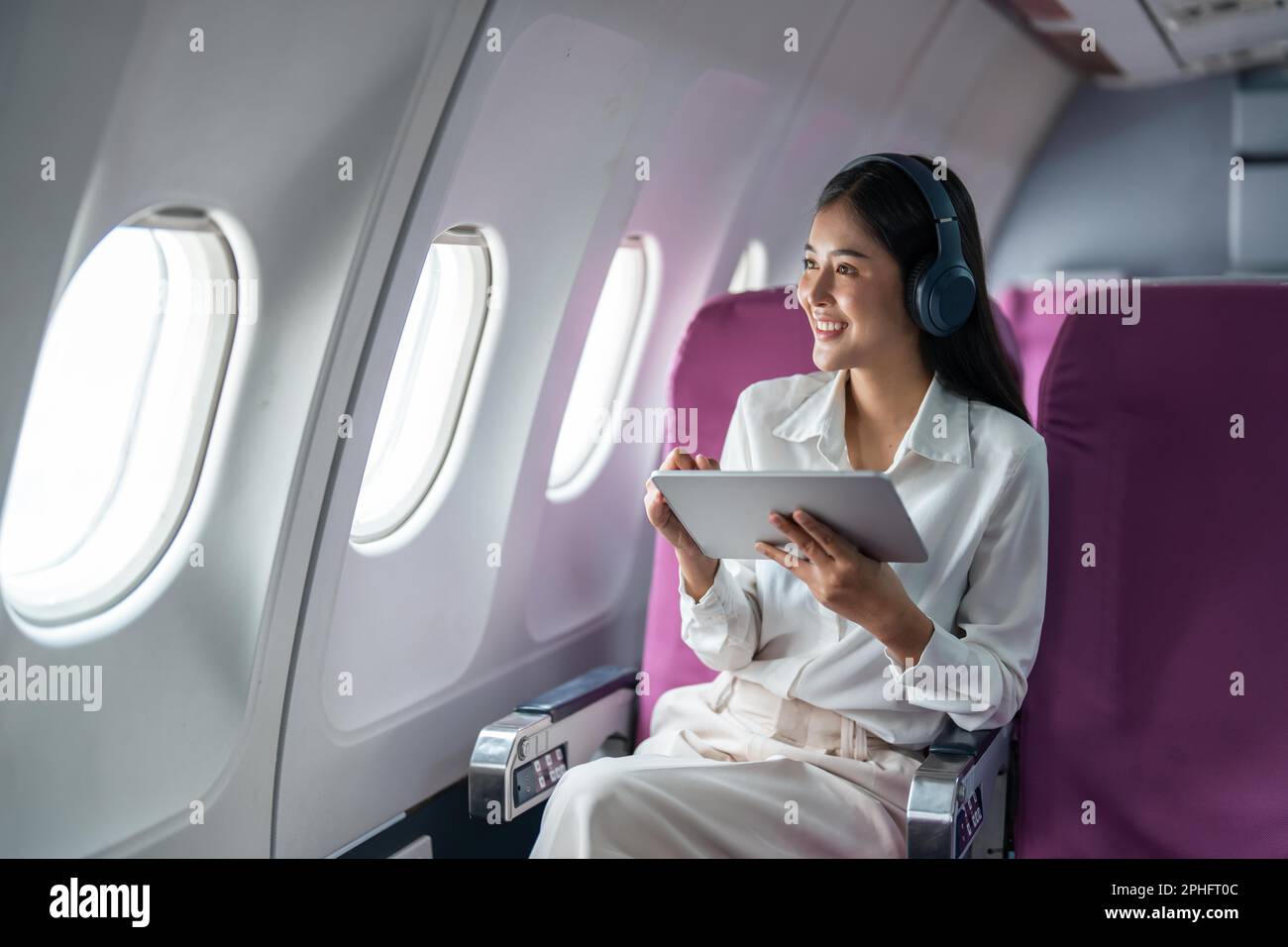 Young Asian business woman or female passenger wearing wireless headphone and working with tablet during the flight Stock Photo