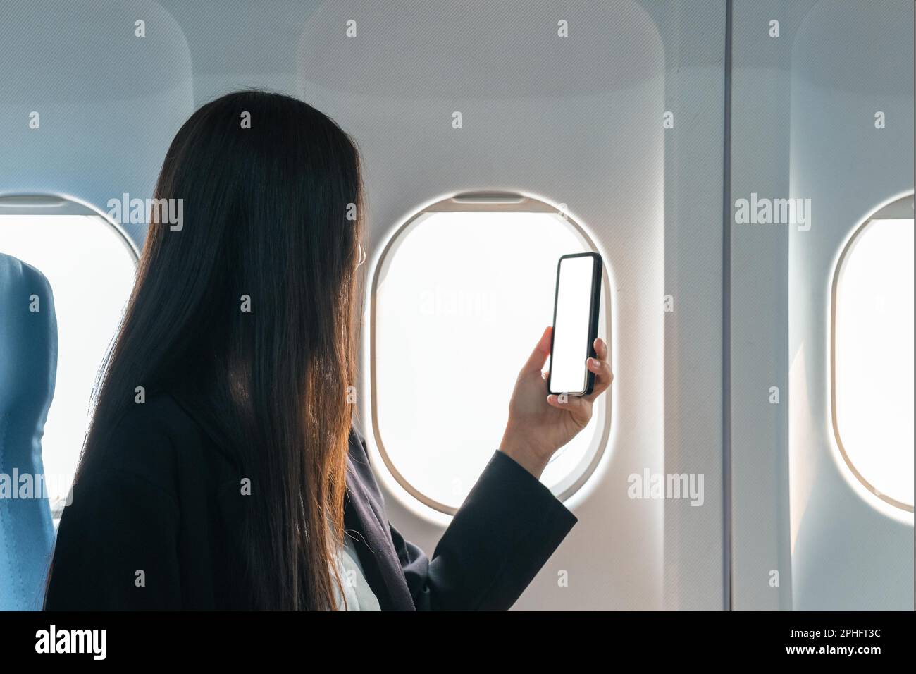 A female traveler passenger sits at the window seat in economy class, using his smartphone, holding a mobile phone white screen mockup. close-up image Stock Photo