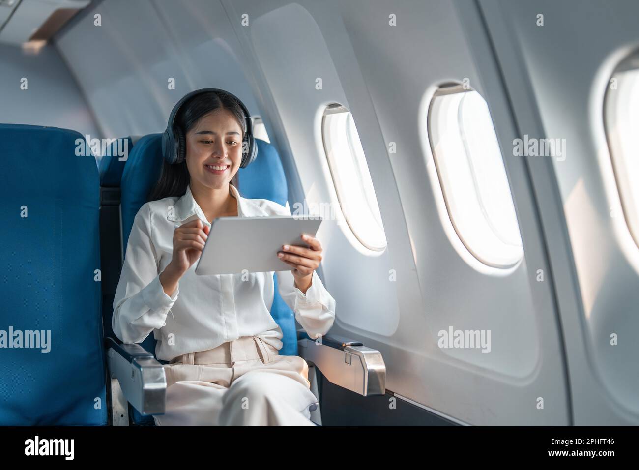 Young Asian business woman or female passenger wearing wireless headphone and working with tablet during the flight Stock Photo