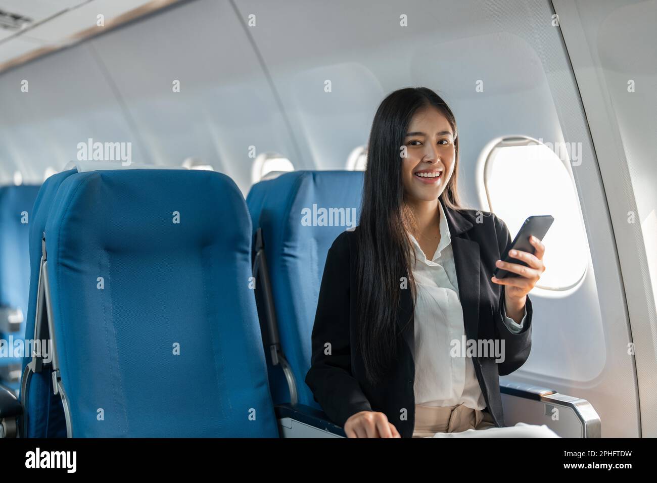 female traveler passenger sits at the window seat in economy class, using his smartphone Stock Photo