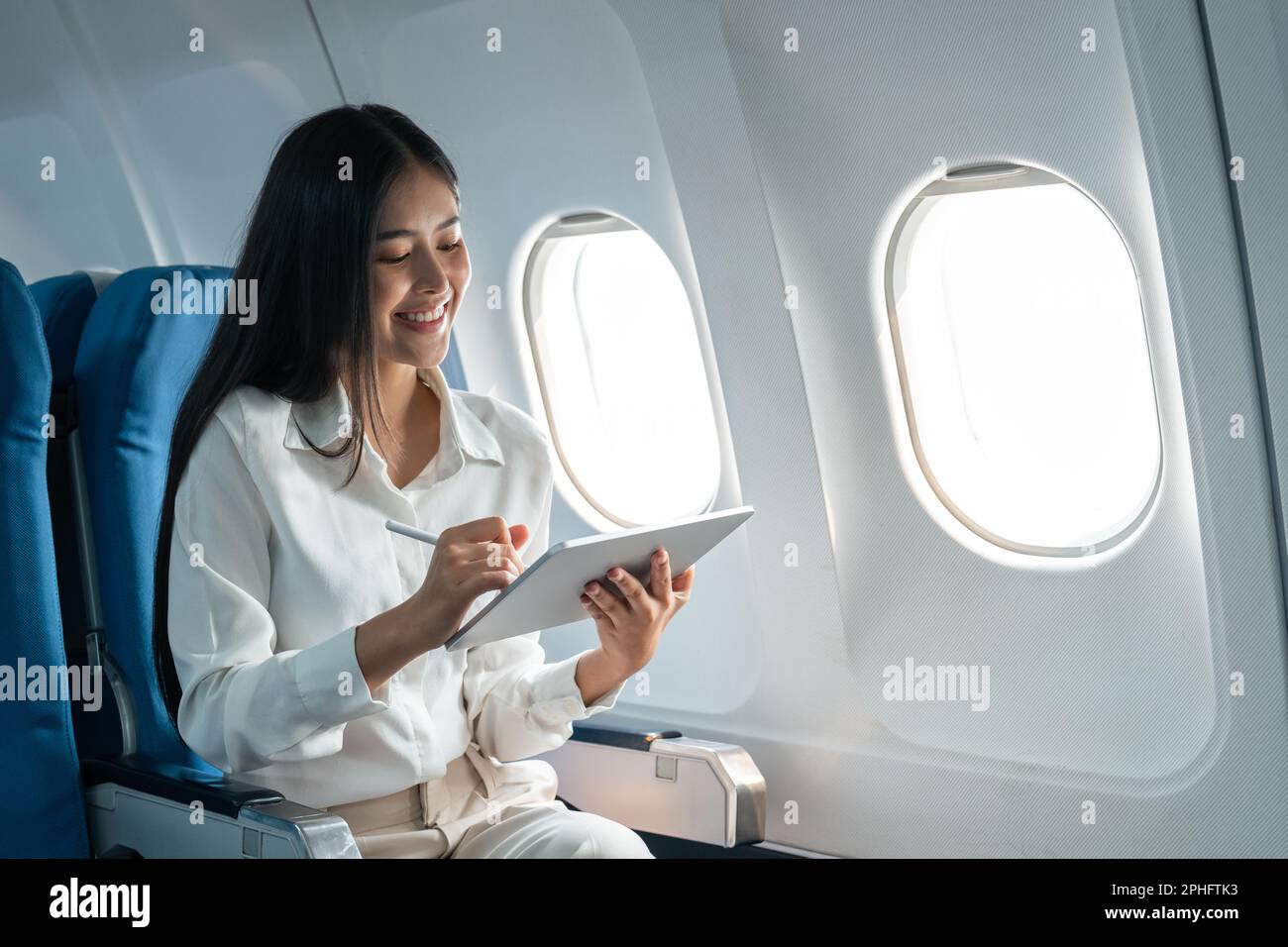 Young asian businesswoman in formal clothes working using tablet with smart pen while sitting in airplane cabin near window traveling to another place Stock Photo