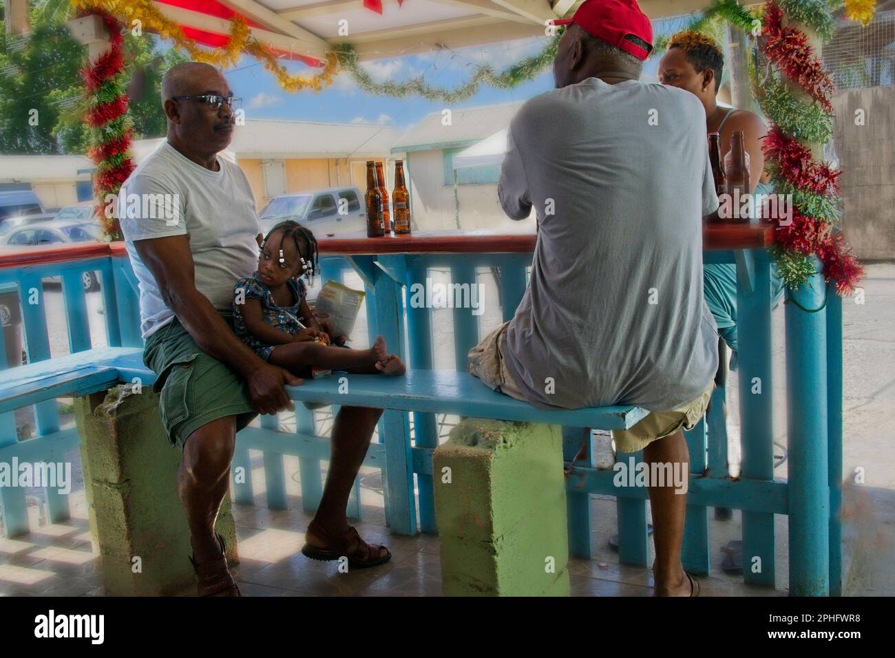 03Feb23 Barbados, Caribbean Three locals and toddler chatting at sunny outside bar Wood seats and colourful decorations Beer bottles on the barrier. Stock Photo