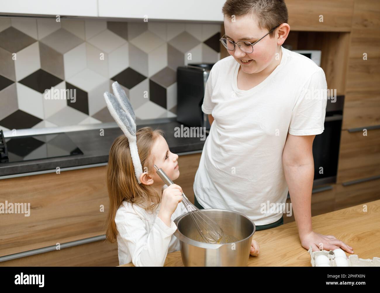 Brother in eyeglasses and his little sister in kitchen smiling and looking at each other while cooking. Child mixing dough in bunny ears. Helping, fam Stock Photo