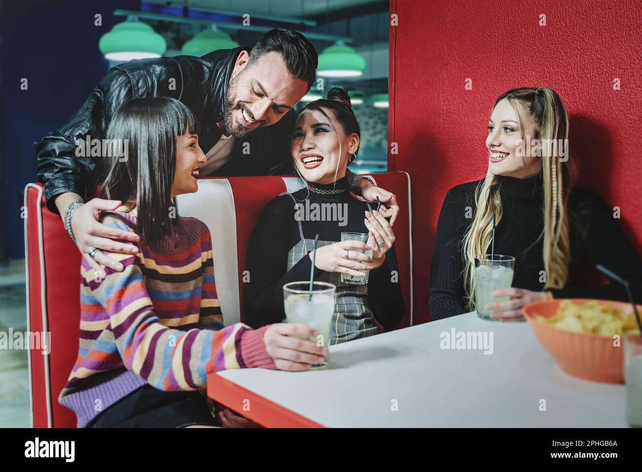 Three female friends enjoy cocktails at a 90s themed game bar, a smiling male friend approaches and hugs two of them, pool tables in the background. Stock Photo
