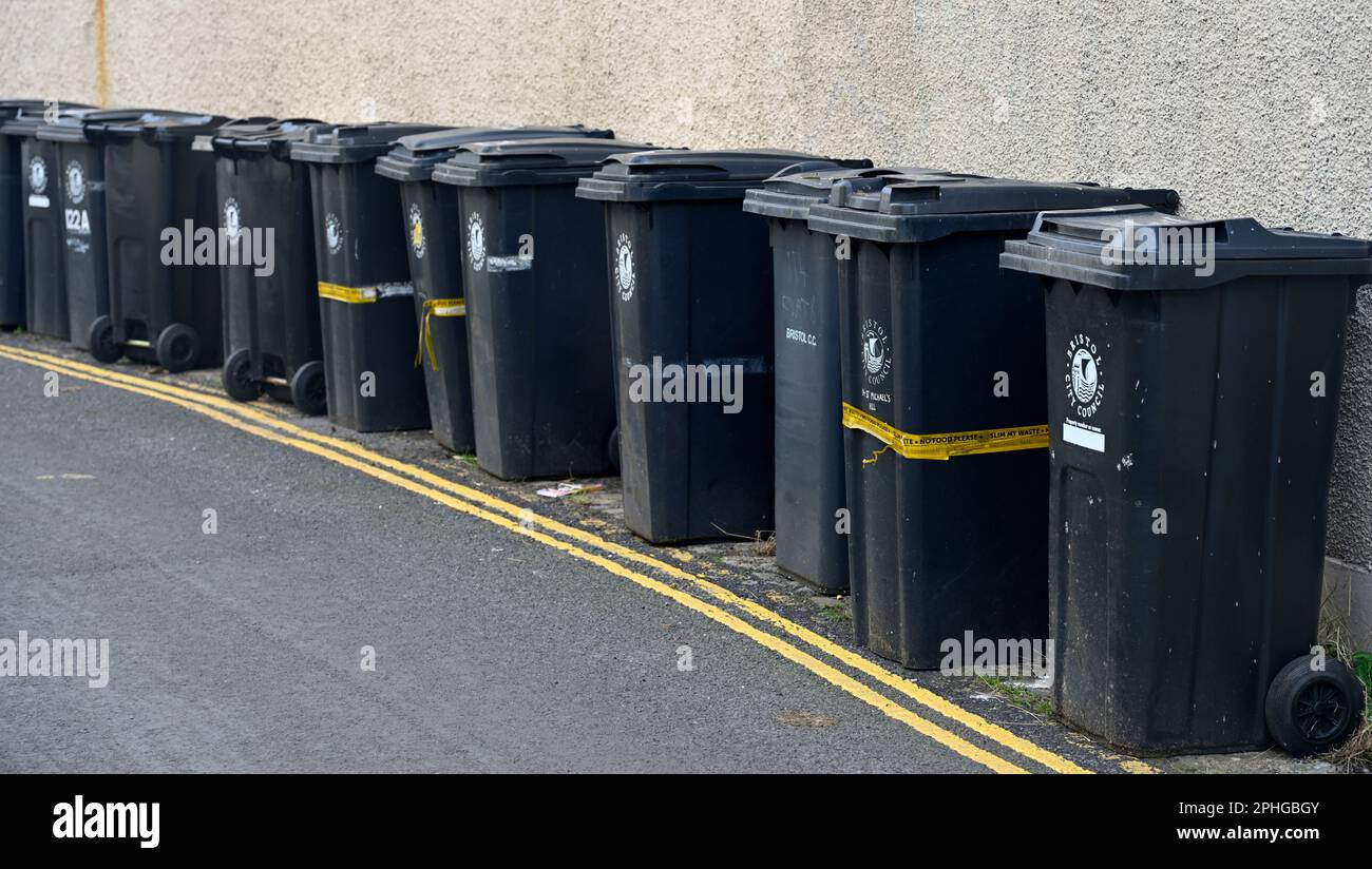 Long row (12) of black domestic rubbish bins out for collection along side of street by house wall, Bristol, UK Stock Photo
