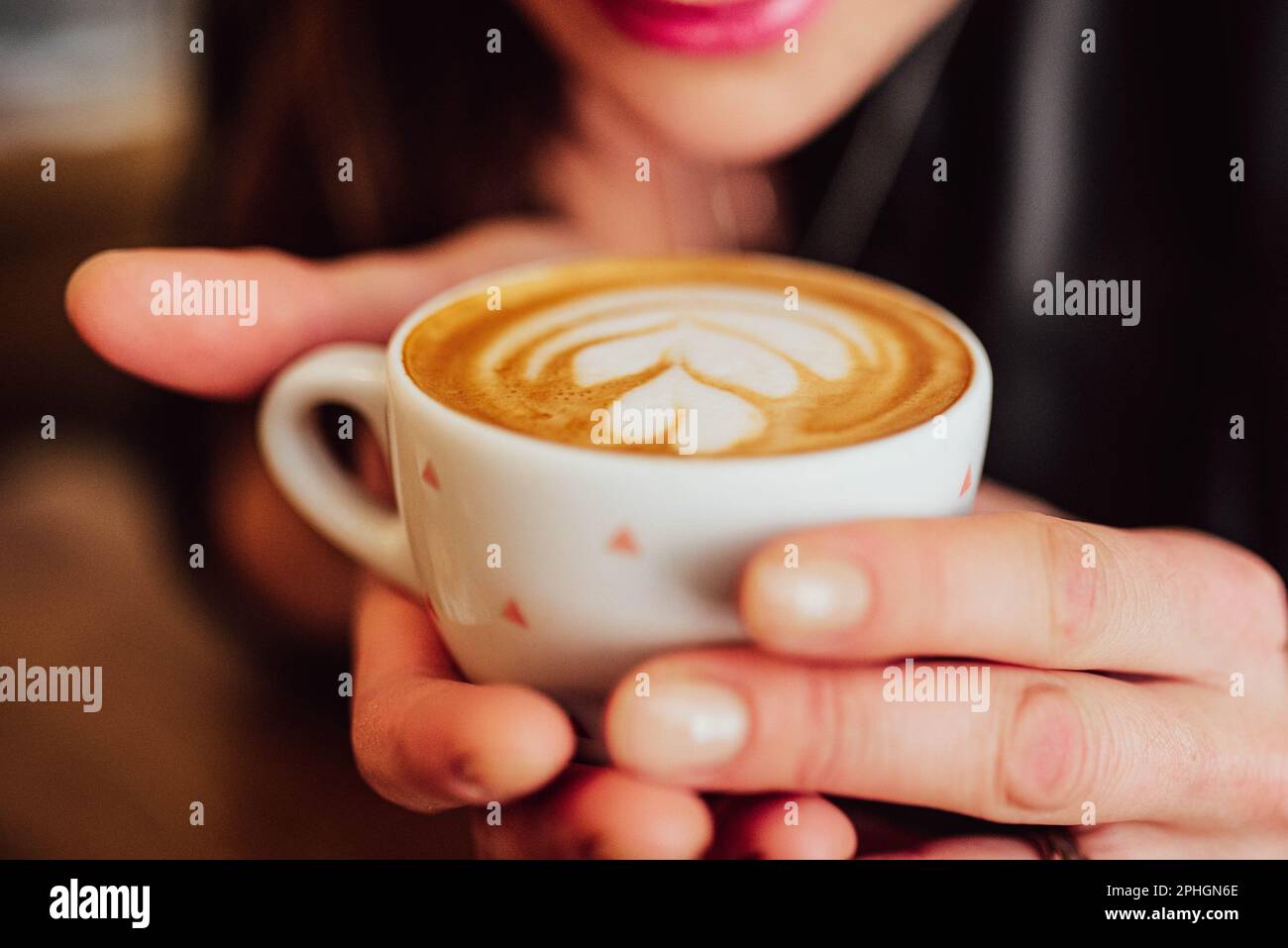 hands holding a white cup with coffee and latte art in a cafe, a woman and painted lips can be seen in the background Stock Photo