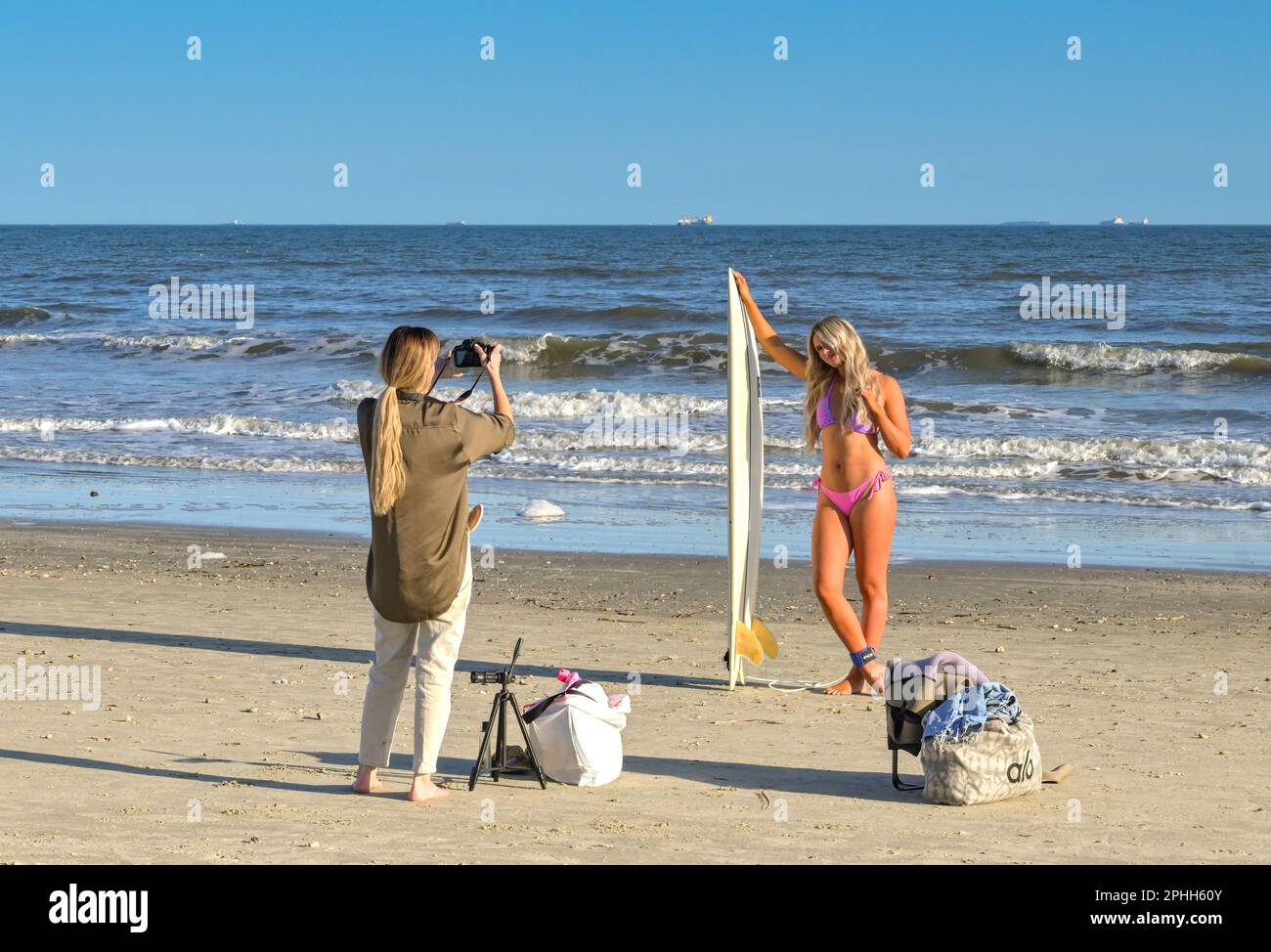 Galveston, Texas, USA - February 2023: Person posing in a swimsuit alongside a surfboard at the water's edge having their picture taken Stock Photo