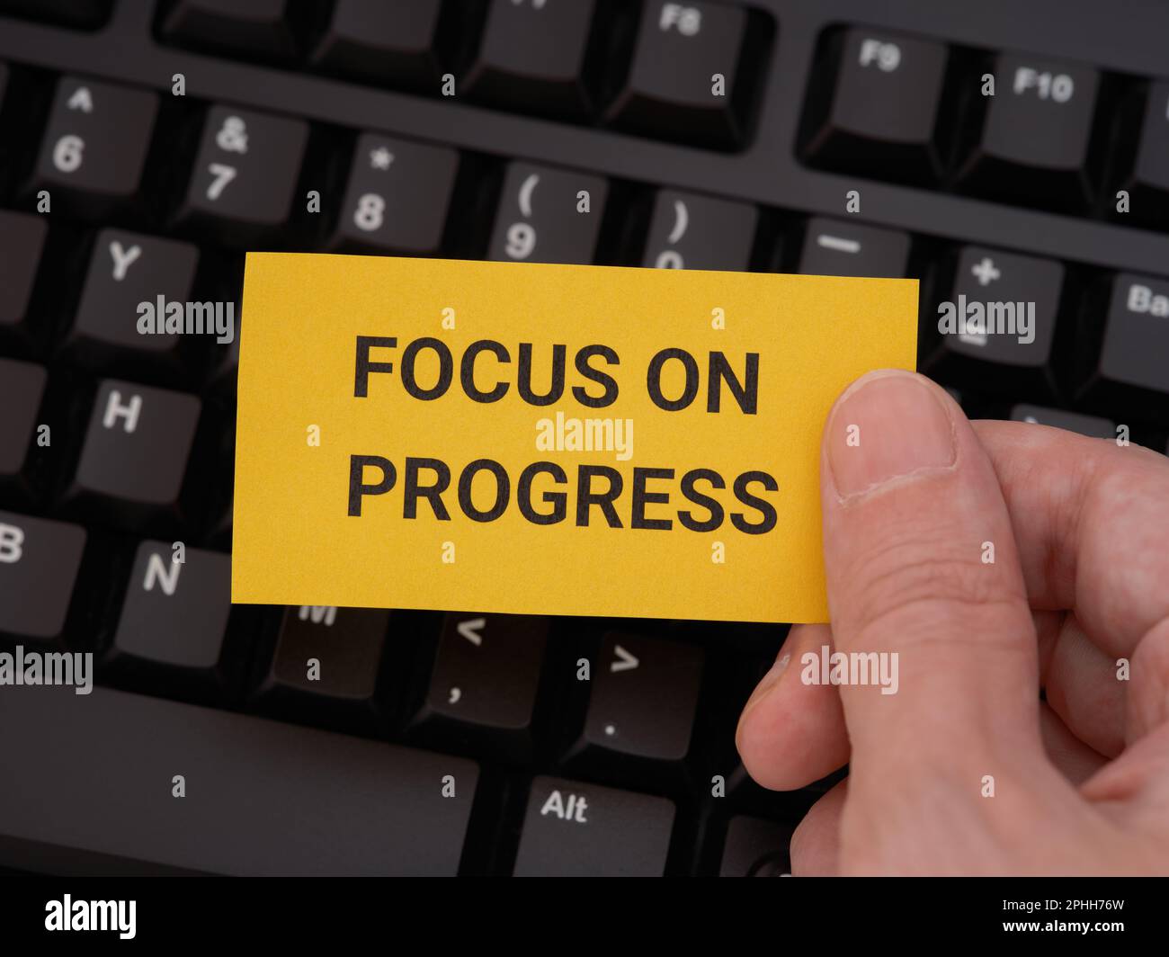 A man holding a yellow paper note with the phrase Focus On Progress on it in his hand against a black computer keyboard. Close up. Stock Photo