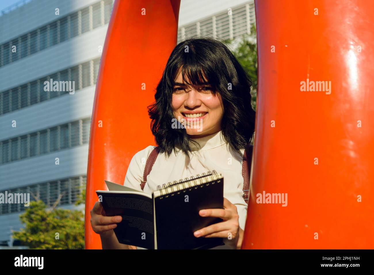young latin woman, short hair and white casual clothes, happy sitting in the college looking at the camera smiling, holding a book in her hands, stude Stock Photo