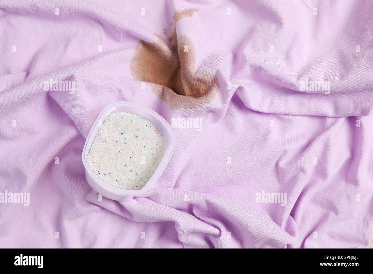 Measuring cup of powdered detergent on garment with stain, above view