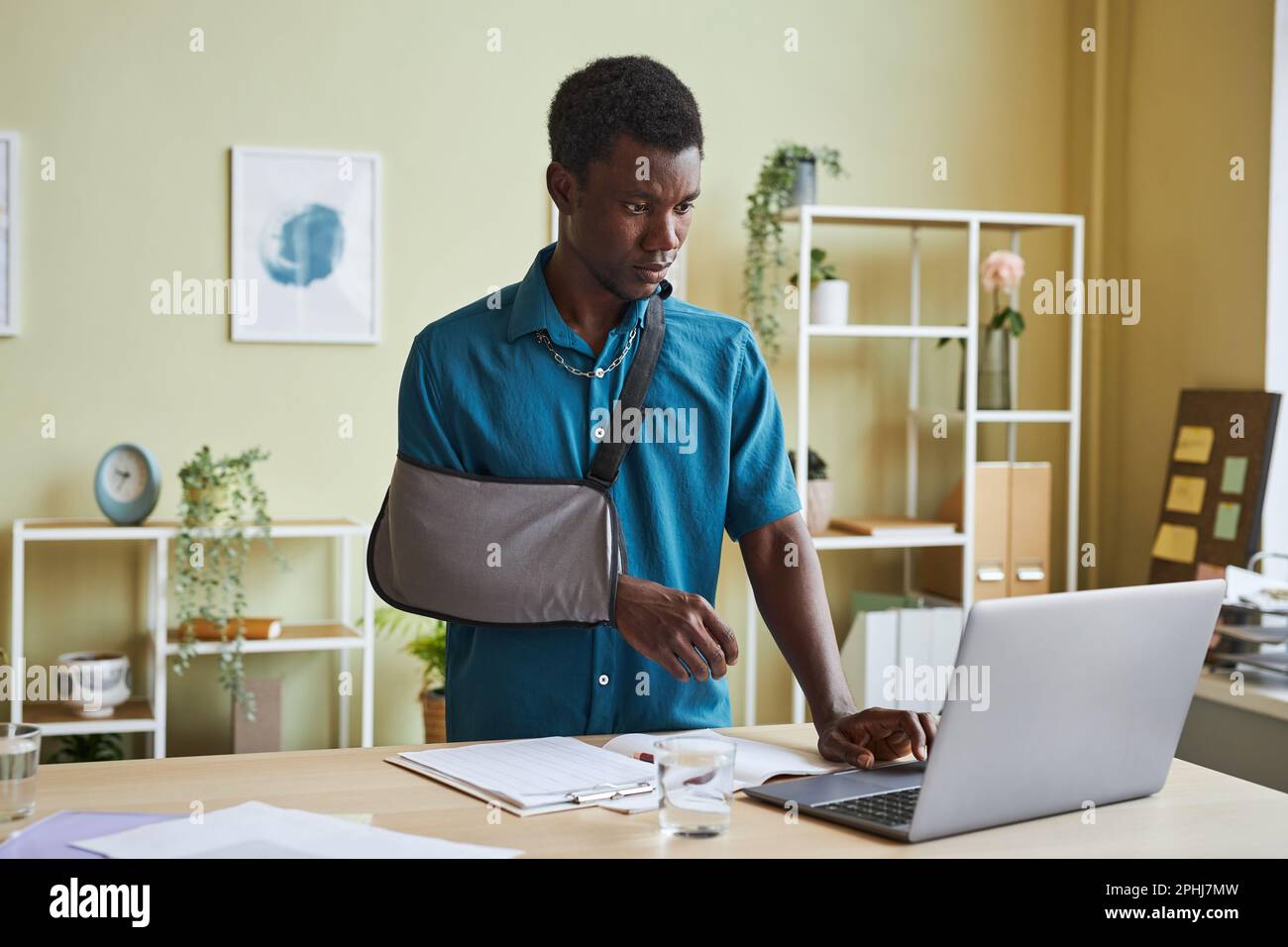 Portrait of black young man with arm sling working at standing desk and using laptop in office Stock Photo