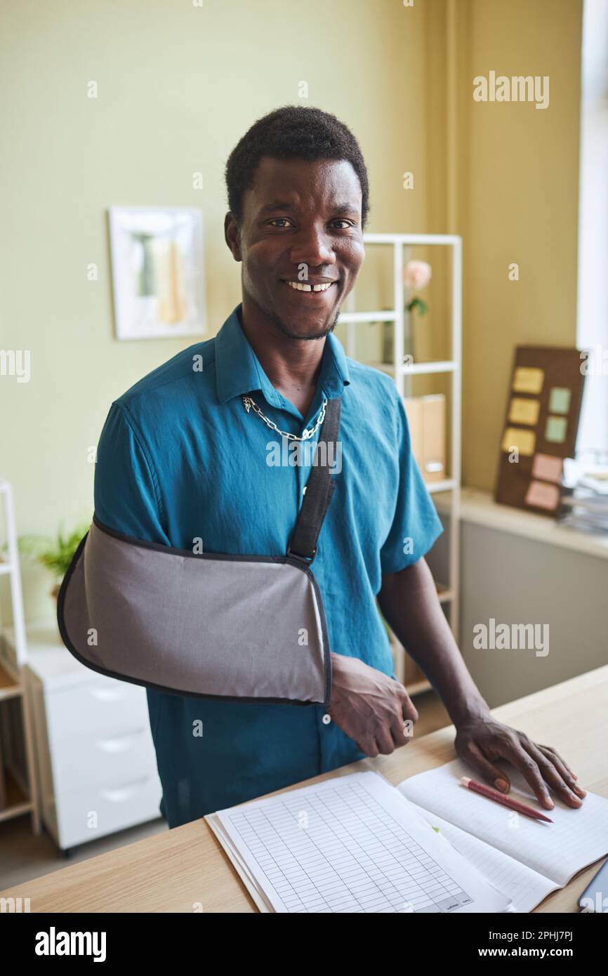 Vertical portrait of black young man with arm sling working at standing desk and smiling at camera Stock Photo