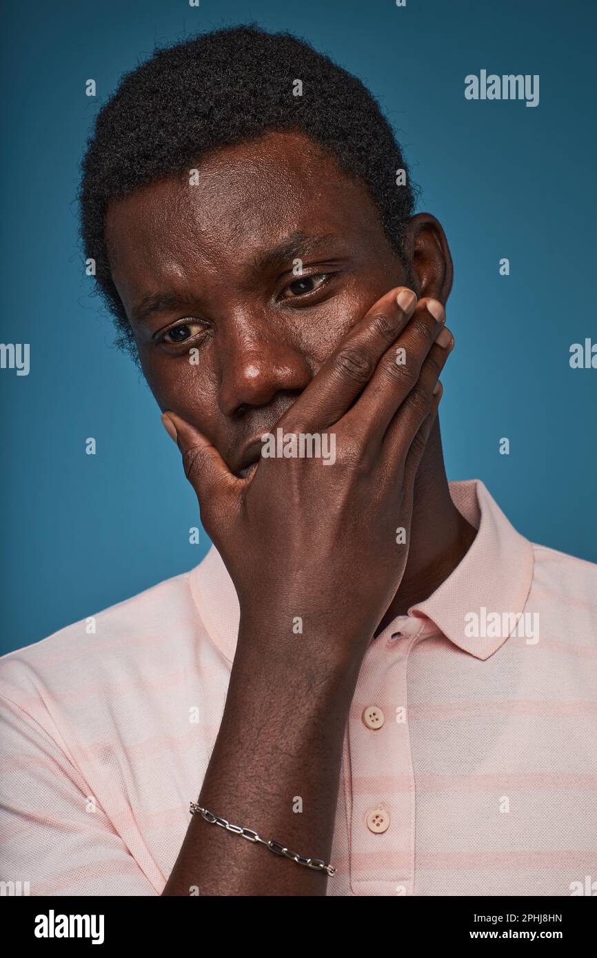 Vertical closeup portrait of distressed black man covering mouth against blue background Stock Photo