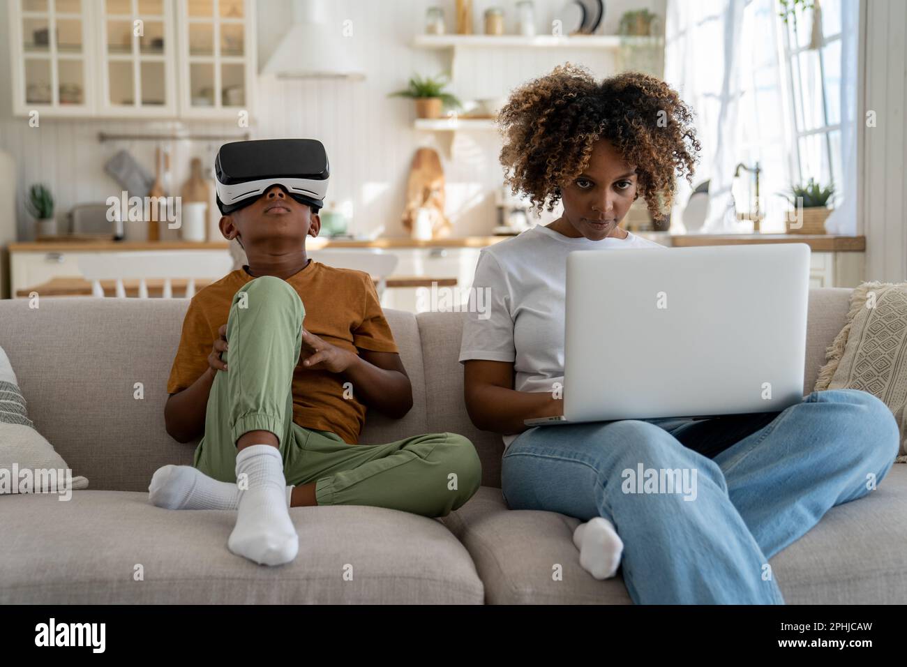 Child using virtual reality simulator headset while sitting on sofa with mother working on laptop Stock Photo