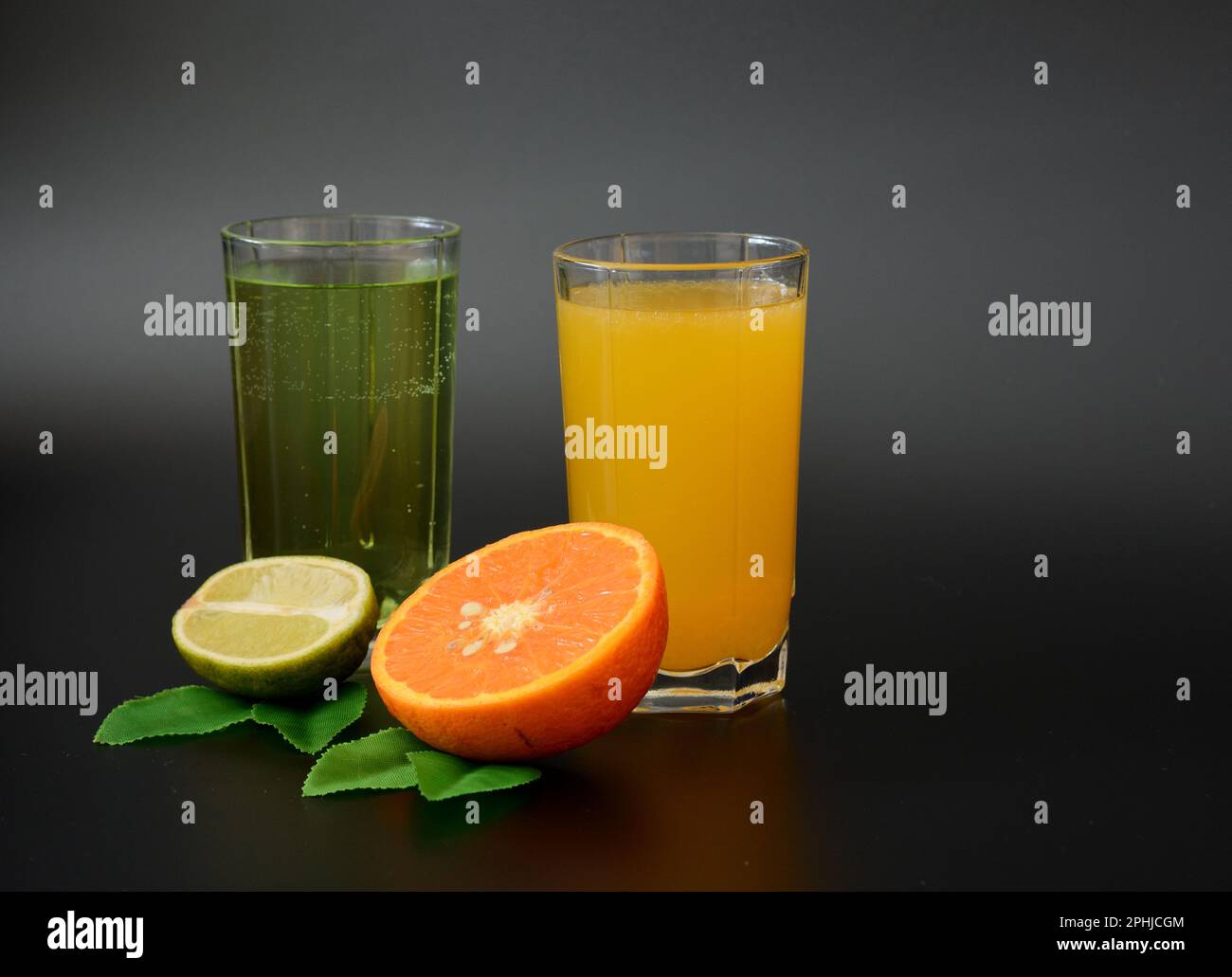 Two glasses with different citrus juices on a black background, next to pieces of ripe orange and lime with leaves. Close-up. Stock Photo