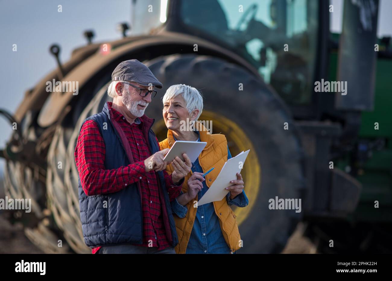 Mature Farmers Man And Woman Standing In Field And Writing Notes While Tractor Plowing In 