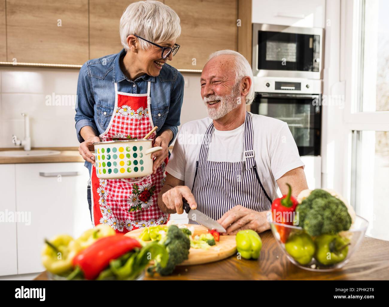 Cheerful senior couple preparing soup, husband cutting vegetable while wife holding pot and smiling Stock Photo