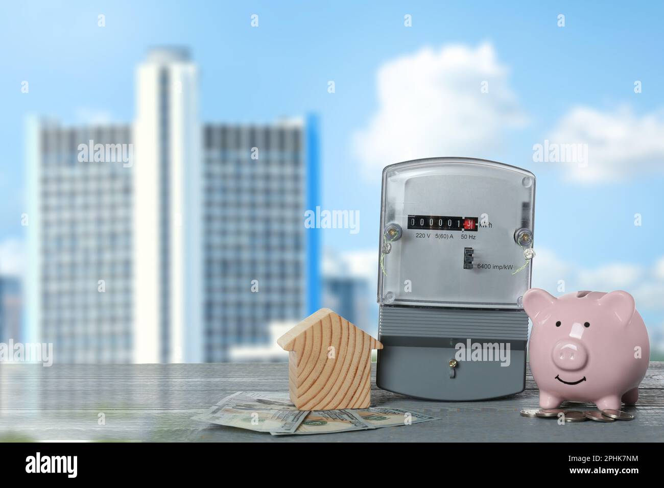 Electricity meter, house model, money and piggy bank on grey wooden table against blurred view of cityscape Stock Photo