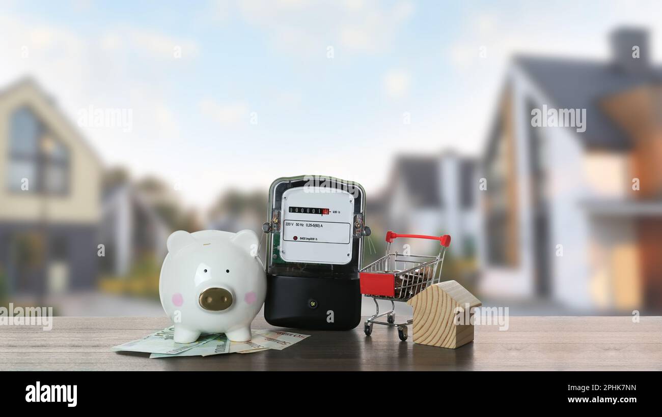 Electricity meter, house model, piggy bank and shopping cart with money on wooden table against blurred view of modern buildings Stock Photo