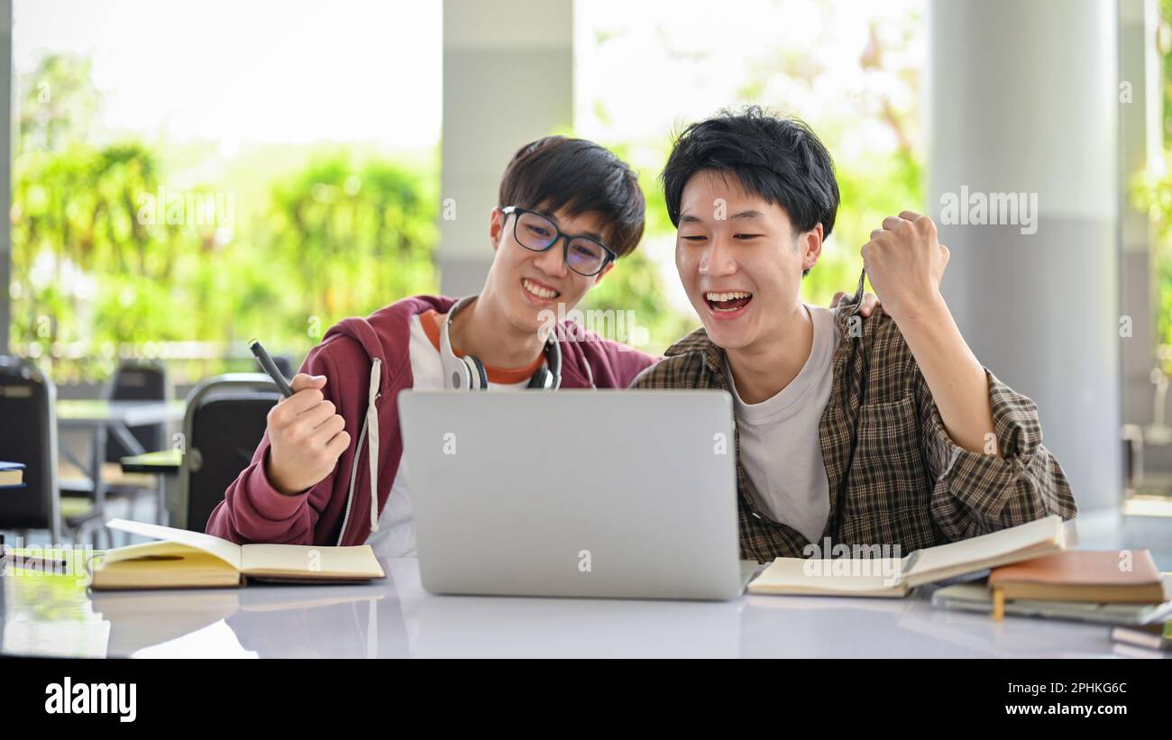 Two smart and cheerful young Asian male college students are looking at laptop screen and celebrating their test score together. Stock Photo