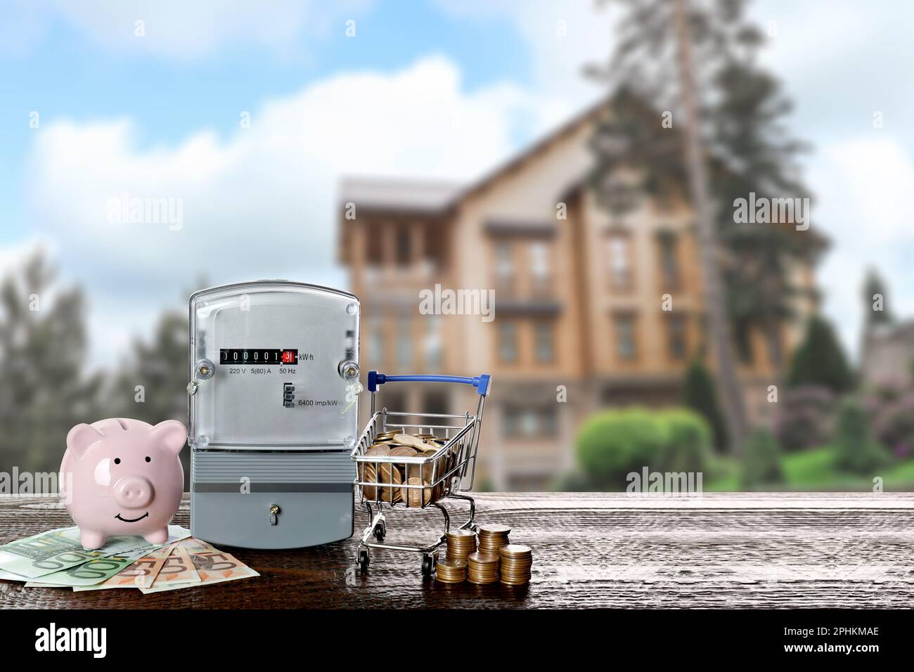 Electricity meter, piggy bank and small shopping cart with money on wooden table against blurred view of beautiful house Stock Photo
