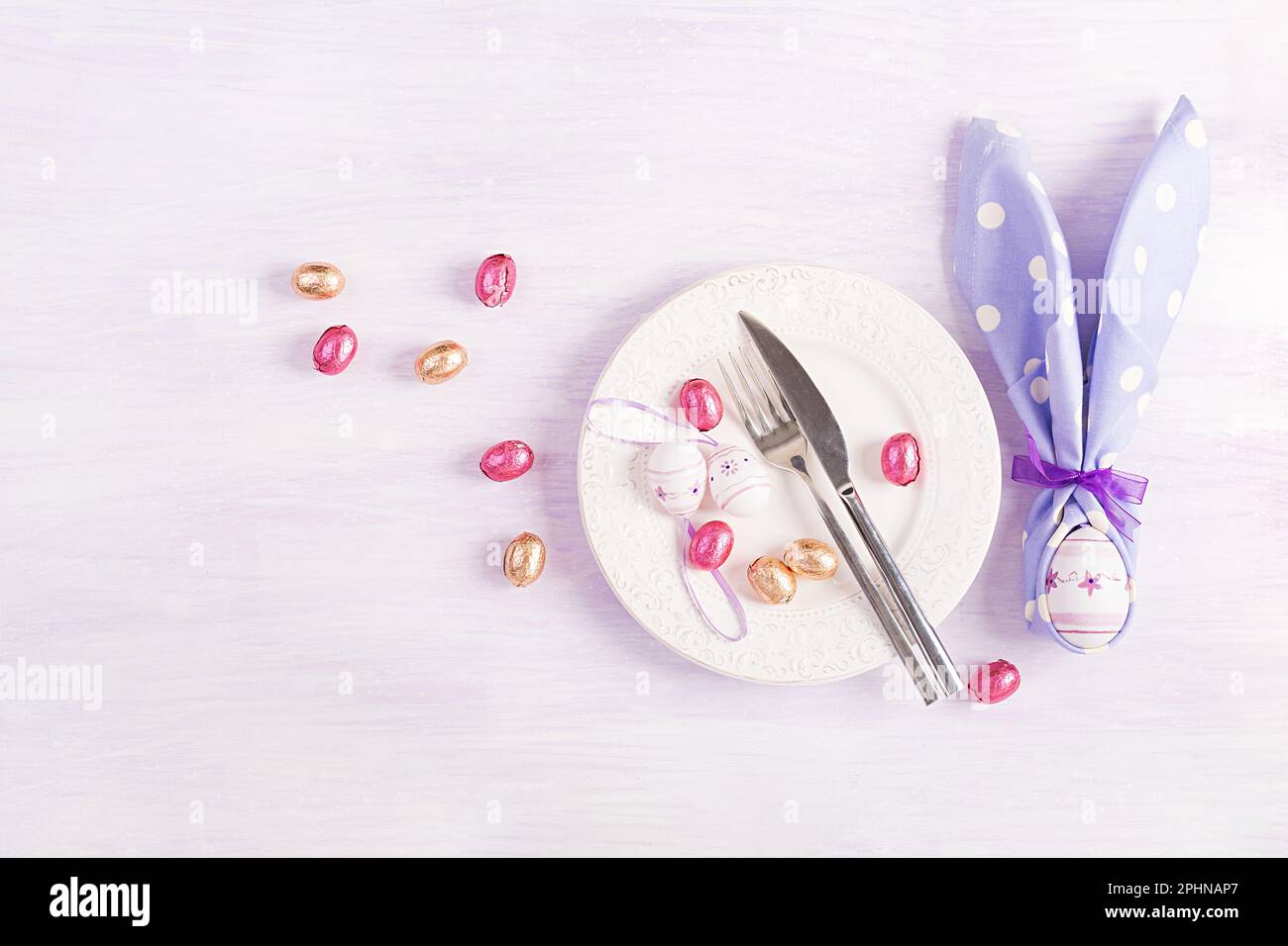 Easter table setting. White plate with a napkin folded in the shape of a rabbit, Easter and chocolate eggs on a pink background. Happy Easter holiday Stock Photo