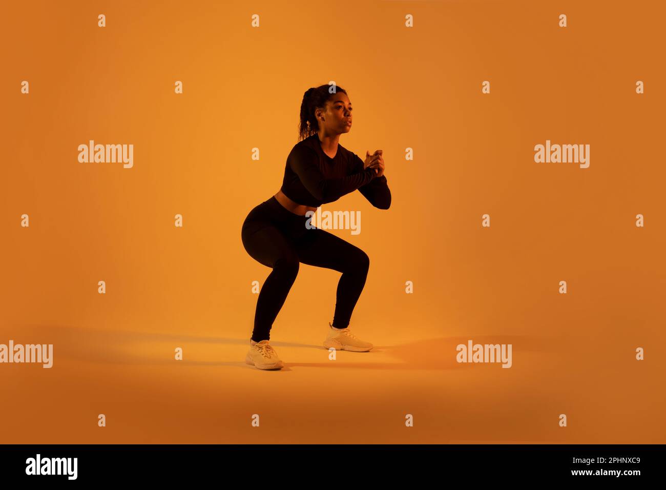 Workout and fitness. Sporty black woman doing deep squat flexing muscles exercising over orange neon background Stock Photo