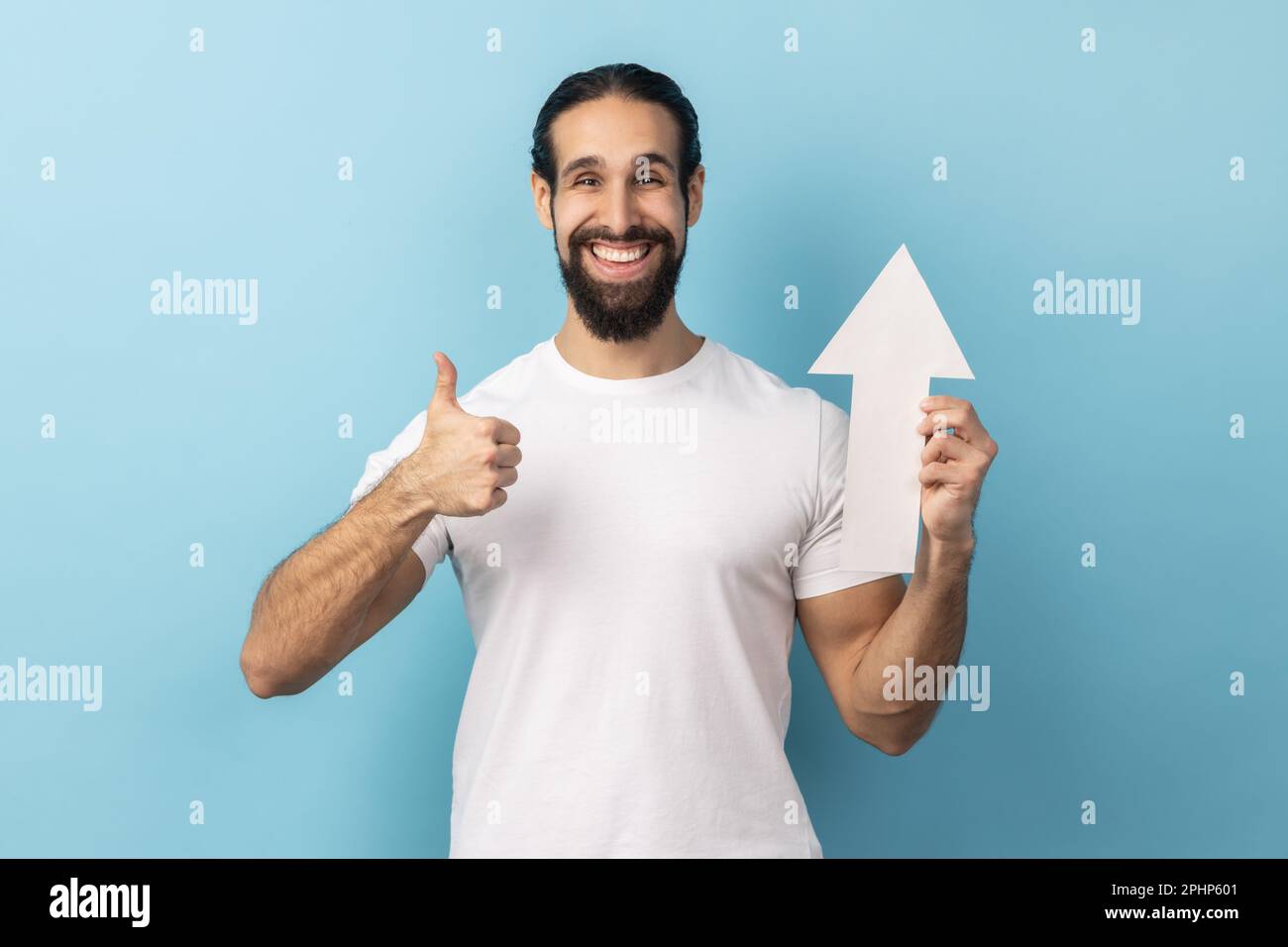 Portrait of happy handsome man with beard wearing white T-shirt showing white arrow pointing up, showing thumb up, like gesture, looking at camera. Indoor studio shot isolated on blue background. Stock Photo