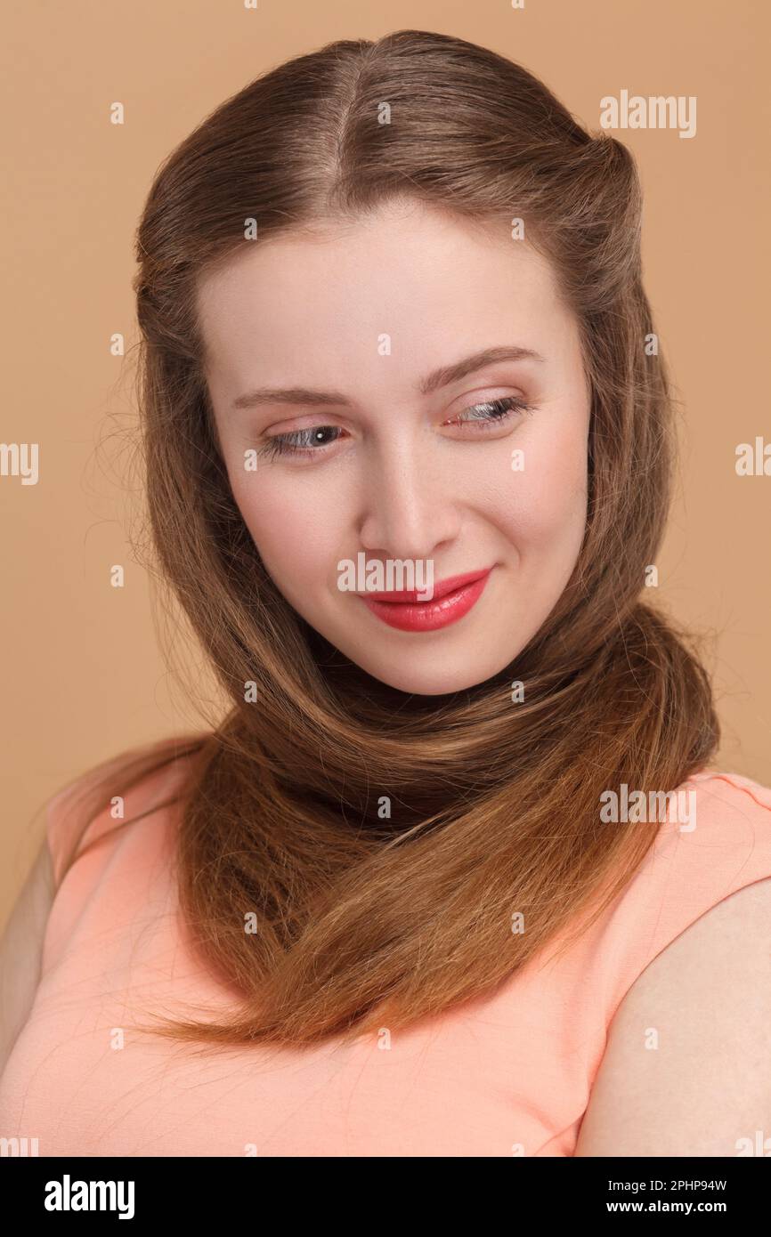 Closeup portrait of beautiful pretty woman with red lips standing wrapped her neck with her long hair, looking away with smile, wearing elegant dress. Indoor studio shot isolated on brown background. Stock Photo