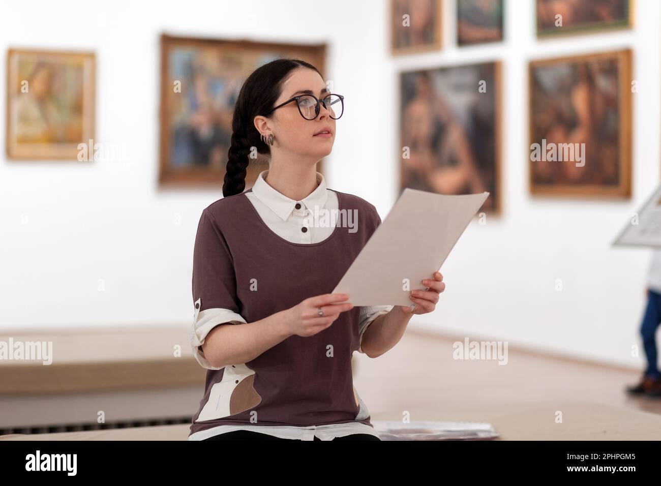 Portrait of caucasian pretty young woman wearing glasses learning educational brochure. Defocused pictures in background. Concept of exhibition in gal Stock Photo