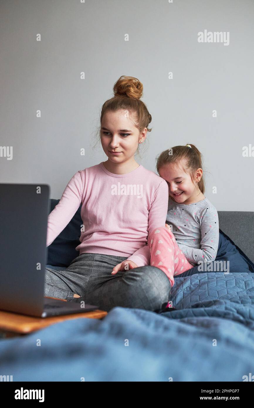 Teenage girl and her younger sister playing learning on laptop spending time together at home during COVID-19 quarantine. Girls sitting on bed in fron Stock Photo