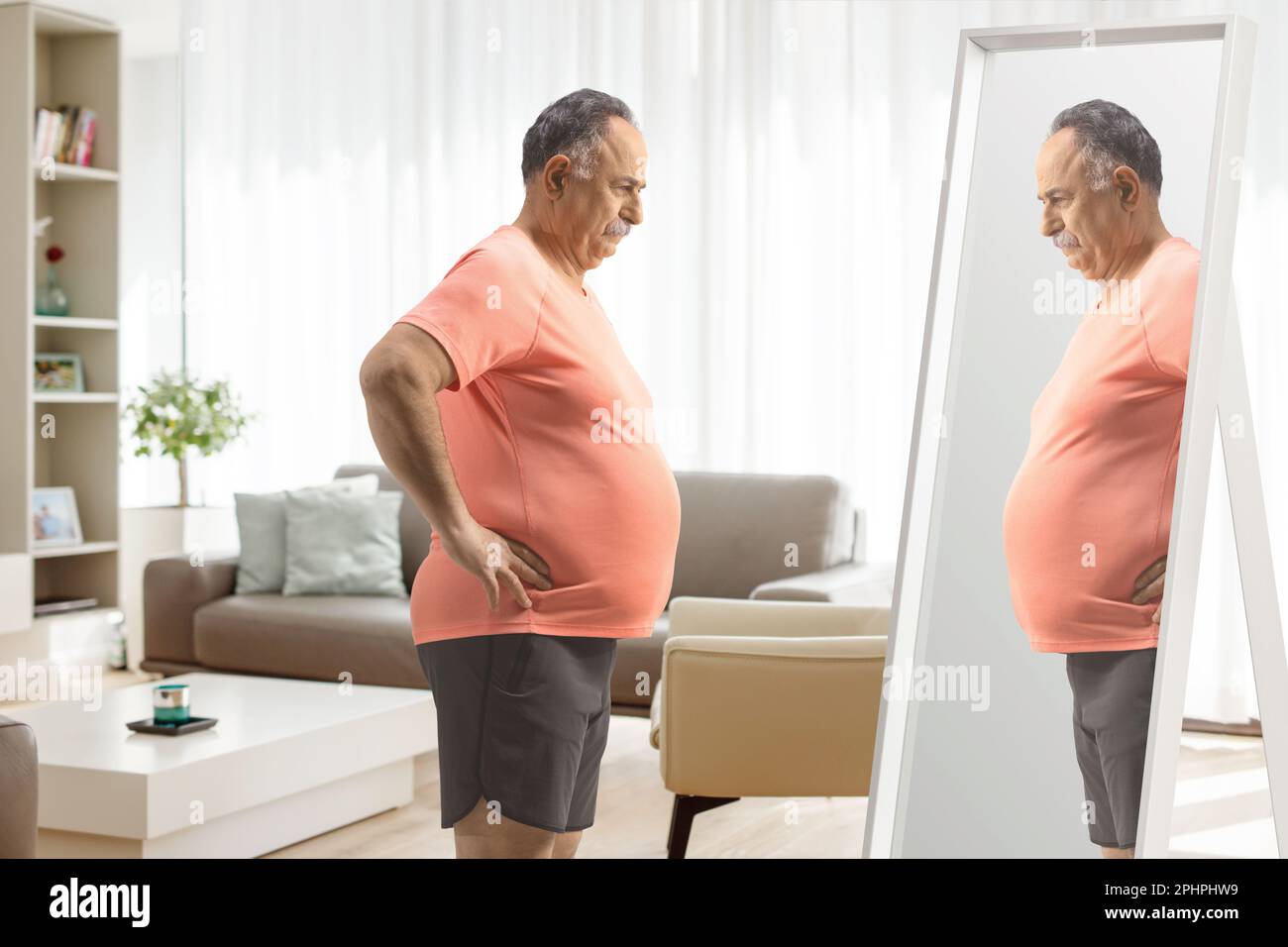 Mature man with a big belly standing in front of a mirror at home in a living room Stock Photo