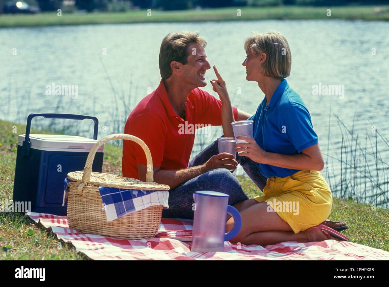 1990 HISTORICAL YOUNG COUPLE SITTING PICNIC ON BLANKET BY WATER Stock Photo
