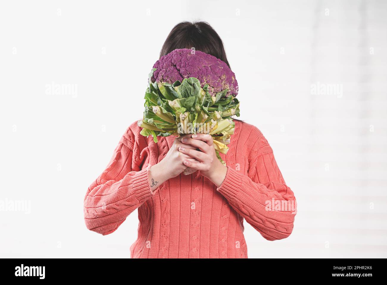 Cheerful young Caucasian woman playfully hiding her face behind a cauliflower, holding it with both hands, standing by a window with a white curtain. Stock Photo