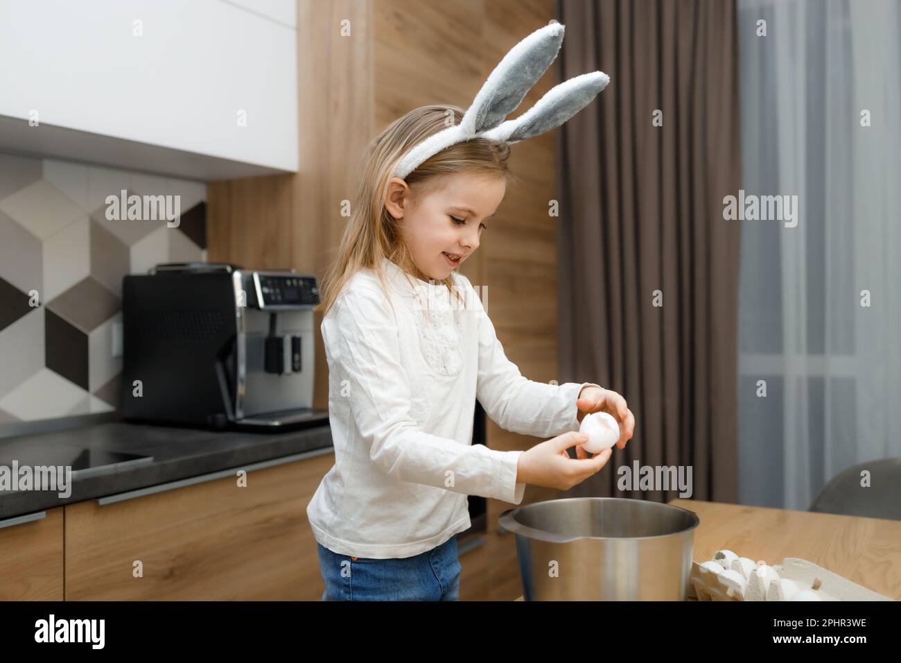 little girl in bunny ears cracking egg into bowl on wooden table in modern kitchen, learning to cook, preparing dough for baking Stock Photo