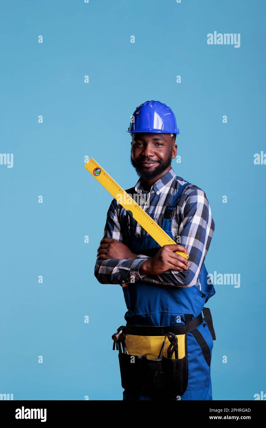 Smiling african american repair worker in uniform holding tape measure on blue studio background. Experienced builder holding work tools wearing coveralls and hard hat. Stock Photo