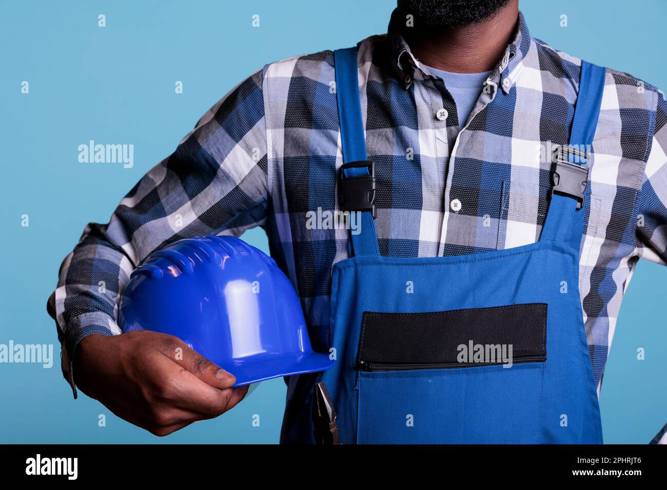 Middle section of construction worker holding protective helmet under his arm. African American man wearing coverall and plaid shirt against blue background, studio shot. Stock Photo