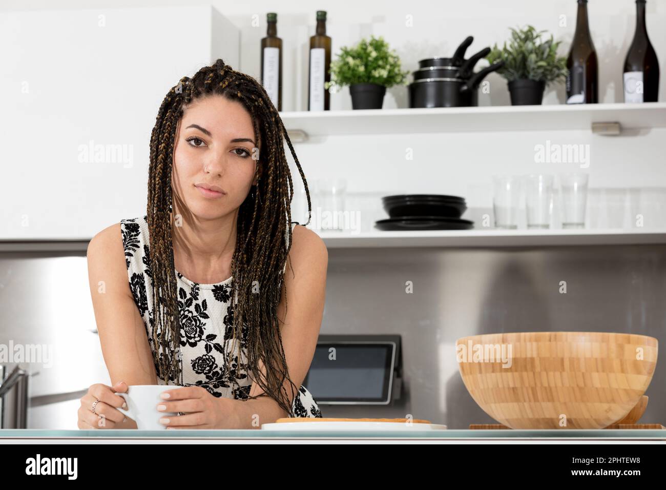 Young woman on a coffee break in a modern kitchen, with an inquisitive and slightly annoyed look. She seems to say that you see only a beautiful girl Stock Photo